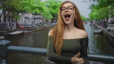 Yeşil omuzlu, yeşil elbiseli bir kadın başparmağıyla kulağına çağrı hareketi yapıyor ve caddedeki bir kanal köprüsünde gülüyor..