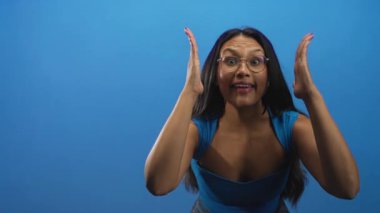 Gözlüklü kadın iki elini de yüzünün iki tarafına kaldırır ağzı açık ve geniş bir bakışla mavi stüdyo arkaplanına bakar; sürpriz merak.