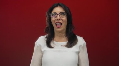 Elleri yanaklarında ve kırmızı stüdyoda ağzı açık, gözlüklü ve beyaz bluzlu bir kadın. Sürpriz..