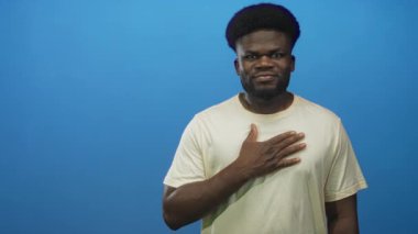 Mavi bir stüdyoda elini göğsüne koyan bir adam, hafif bir gülümseme ve bir söz jesti göstererek başını eğdi; sessiz bir minnettarlık.