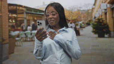 Göğüs göğüse akıllı telefon tutan, kaşları çatık, gözlük takan, sokakta tepki veren bir kadın, endişe verici bir bağlantı..