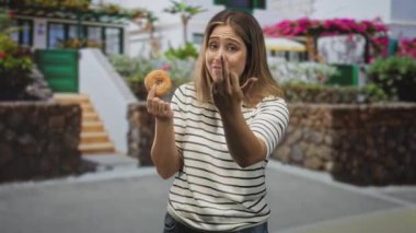 Çizgili tişörtlü kadın bir çörek tutuyor ve eli kameraya doğru bir güneş ışığı caddesinde işaret ediyor; neşeli bir davet.