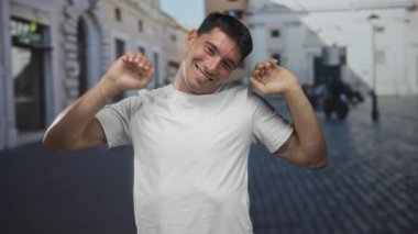 Güler yüzlü İspanyol adam gündüz vakti şehir meydanındaki güneş ışığıyla aydınlanan kaldırım taşı caddesinde yanak jesti yapıyor..