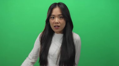 Uzun saçlı bir kadın, izole edilmiş yeşil arka planda beyaz gömlek giyerek şaşırmış bir ifade sergiliyor.