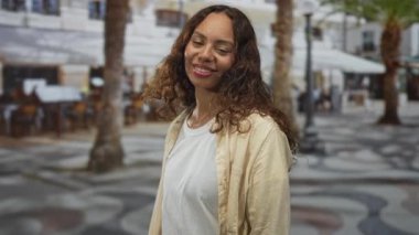 Güneşli bir sokak meydanında, palmiye ağaçları, açık kafe koltukları ve kaldırım taşı desenli bir kameraya bakan bir kadın..