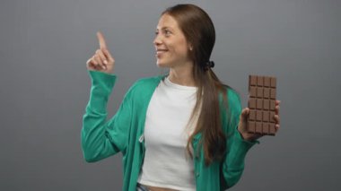 Büyük bir çikolata tutan ve gri stüdyoda yeşil hırka ve beyaz bluz giyen bir kadın. Neşeli..