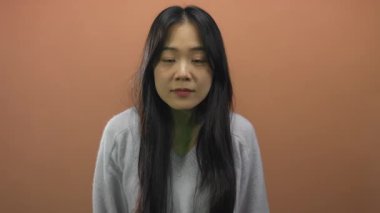 Genç Çinli bir kadın, izole edilmiş kahverengi arkaplan duvarı üzerinde şaşkınlığını dile getiriyor. Duygularını, geniş gözlü ve elleri yüzlerinde, rahat bir süveter içinde, sade bir arka fona doğru vurguluyor..