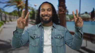 Adam iki eliyle sokakta su kenarında barış işareti yapıyor. Kot ceket ve beyaz tişört giyen palmiye ağaçları. Huzurlu neşe..