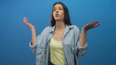 Young hispanic woman shrugging with raised hands in a studio against a bright blue backdrop, wearing denim overshirt and yellow tank top, palms up; confusion.