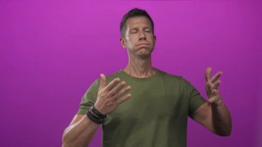 Elleri ve bilezikleriyle hareket eden, mor stüdyoda yeşil tişört giyen beyaz bir genç..