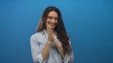 Genç İspanyol kadın mavi stüdyoda çıplak ellerini çırpıyor geniş bir gülümseme ve rahat bir duruş ile; neşe.