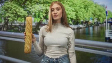 Kanal parmaklıklarında, sol eli ve sağ eliyle baget tutan kızıl saçlı kadın, kot pantolon ve balıkçı yaka giyiyor; eğlenceli bir merak..