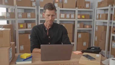 Masada dizüstü bilgisayar ve paketle oturan bir adam, kollar birleştirilmiş ve raflar ve karton kutular görülebilir durumda; güven verimliliği.