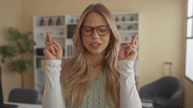 Gözlüklü kadın modern ofiste şans umuduyla masa başında parmaklarını çaprazlıyor..