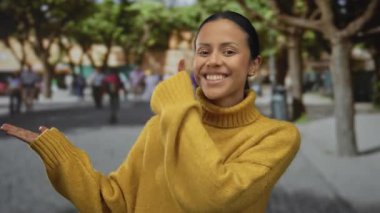 Sarı süveter giyen kadın ağaçlar ve arka plandaki insanlarla dolu caddede neşeyle gülümsüyor..