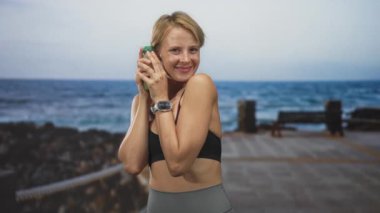 Kadının elinde yeşil meyve suyu şişesi var. Spor sutyeniyle çıplak göbek ve akıllı kaslarını gösteren bir iskelede. İçeceği incelerken gülümsüyor..