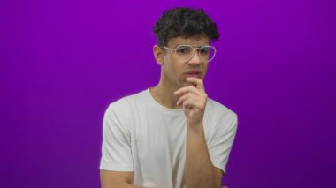 Gözlüklü ve eli çenesinde olan bir adam stüdyo ortamında beyaz tişört giyerek poz veriyor; belirsizlik.