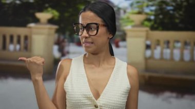 Kadın dışarıda baş parmağıyla işaret ediyor, taş bir balkonun önünde duruyor, gözlük takıyor ve beyaz bir bluz giyiyor, samimi ve düşünceli bir anı yakalıyor..