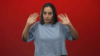 Avuç içleri kalkık, elleri öne dönük genç İspanyol bir kadın, tarafsız bir ifade, kırmızı arka planı olan bir stüdyoda günlük tişört giyiyor, teslim oluyor..