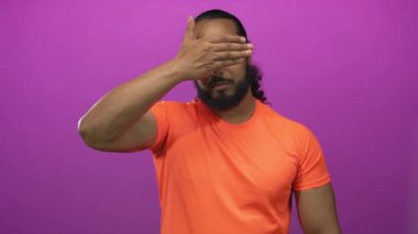 Adam gözlerini sağ eliyle kapatıyor ve turuncu tişört giyerken mor bir stüdyoda gülümsüyor..
