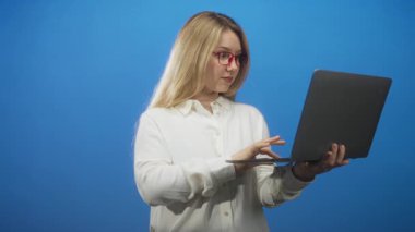 Dizüstü bilgisayarı sol eliyle tutan ve sağ eliyle tuşlara basan bir kadın mavi bir stüdyo arkaplanına karşı kırmızı gözlük takarken; profesyonel özgüven.