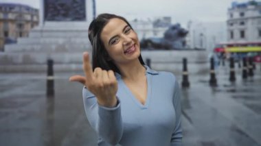 Şehir caddesinde gülümseyen bir kadın, açık havada, ikonik simgelerle ve açık mavi bluz giyen yağmurlu gün atmosferiyle davetkar bir jest yapıyor..