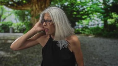 Büyük bir ağacın yanındaki yeşil bir parkta el pençe divan duran gözlüklü kadın, irkilerek ve sesten başını çeviriyor; rahatsız edici sesler duyuyor.