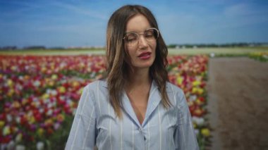 Gözlüklü ve mavi bluzlu kadın orman açıklığında renkli lale çiçeklerinin arasında gülümsüyor..