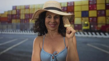 Mayolu ve şapkalı kadın, arka planda nakliye konteynırları olan bir limanda dondurma yiyor. Boş zamanları ve yaz havasını simgeliyor..