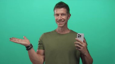 Akıllı telefonu tutan ve yeşil stüdyoda kameraya avucunu açan adam; dost canlısı ürün tanıtımı.