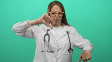 Steteskoplu genç İspanyol kadın doktor turkuaz stüdyoda baş parmağını kaldırıyor, profesyonel olarak onaylamıyor..