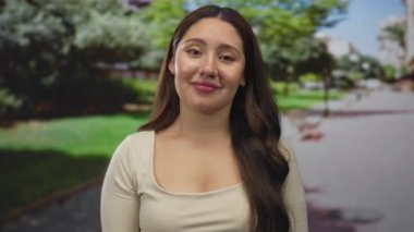 Çıplak köprücük kemiği ve dalgalı uzun saçları olan bir kadın, güneşli yeşil bir parkta gülümserken parmak uçlarına dolaşır ve düşünür..