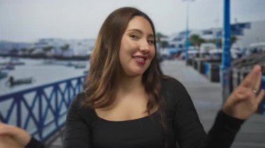 Siyah giysili, gülümseyen yüz dalgaları takınan bir kadın sokakta beyaz bir teknenin yanında el sallıyor..