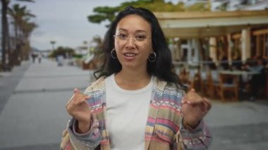 Gözlüklü ve desenli hırka giyen bir kadın. Elleri açık gülümsüyor, avuç içleri açık. Sokakta kameraya doğru hareket ediyor..