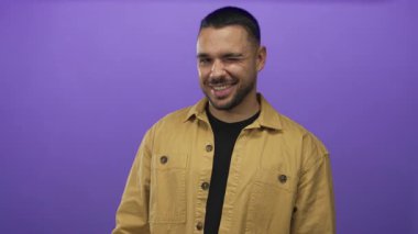 Mor bir stüdyoda, yüzünde sakallı bir gülümsemeyle gülümseyen bir hardal tişörtü giyen İspanyol genç adam..