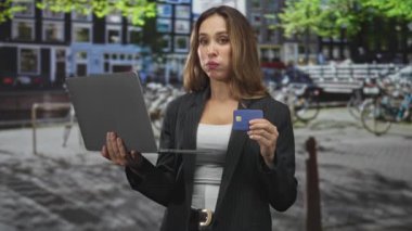 Kadın dizüstü bilgisayar tutuyor ve sokakta bisiklet ve kanal binaları arasında kredi kartını gösteriyor, spor ceket ve beyaz bluz giyiyor; kendine güvenen ticaret.