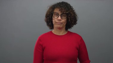 Mature hispanic woman wearing red sweater and eyeglasses makes gesture with hand in studio gray wall; peace harmony unity.