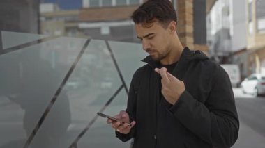 Güneşli bir caddede akıllı telefonu kontrol eden genç İspanyol adam dışarıda heyecan ve enerji gösteriyor..
