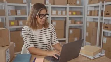 Dizüstü bilgisayarda elleri klavyeli bir kadın gözlüklü ve çizgili bir şekilde bir binada istiflenmiş nakliye kutuları arasında yazı yazıyor; küçük işletmelere odaklan.