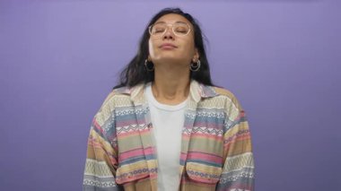 Gözlüklü ve desenli ceketli bir kadın başını arkaya eğiyor, gözleri kapalı stüdyoda mor bir duvarla; sakin tefekkür.