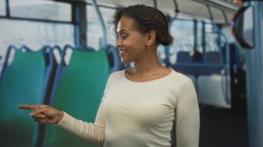Kadınlar, otobüsün içindeki yeşil otobüs koltuğunu gösterirken, aynı zamanda gülümsüyor ve krem rengi bluz giyiyor..