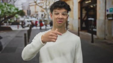 Çağrı jesti olarak kaldırılan İspanyol adam bulanık yayaların ortasında hareketli şehir sokağında gülümsüyor; sosyal bağlantı.