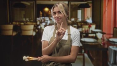 Restoranda sessizlik için işaret parmağıyla dudaklara fırça tutan genç sarışın makyöz; huzur.