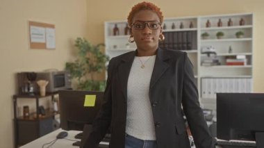 Gözlük takan ve kollarını çapraz bağlayan küpe takan bir kadın bir binada bilgisayar monitörünün yanındaki masaya yaslanıyor..