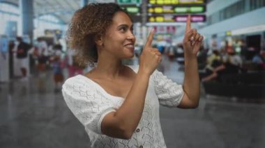 Kadın, gülümseyerek ve yukarı bakarken kalkış kapısının yanındaki yoğun havaalanı terminalinde hareket tahtasını işaret ediyor; sevinçle seyahat.