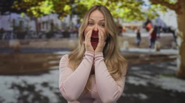 Sarışın kadın, elleri ağızına doluşmuş, sokak meydanında bağırıyor, coşku ve enerji ifadesi..