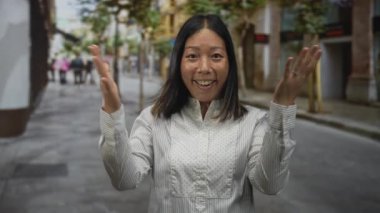 Kadın her iki elini kaldırır çıplak eller ve bir gülümseme göstererek, beyaz çizgili bluz giyerek kalabalık bir şehir caddesinde ağaçlar ve binalarla dururken; memnuniyet;.