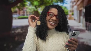 Kıvırcık saçlı ve kırmızı gözlüklü bir kadın ev anahtarlığı ve akıllı telefonu işlek şehir caddesinde tutuyor; ev sahipliği keyfi.