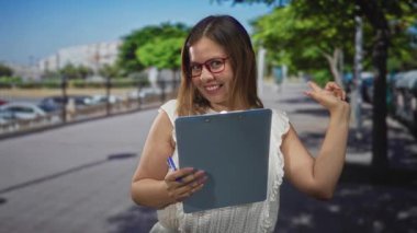 Kırmızı gözlüklü ve beyaz bluzlu kadın elinde pano ve kalem parmağıyla caddeyi işaret ediyor; hazırlık güveni planlıyor.