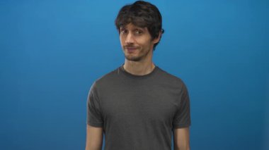 Genç adam tesadüfen gülümsüyor ve parlak mavi bir stüdyoda iki parmağını kameraya doğrultuyor..